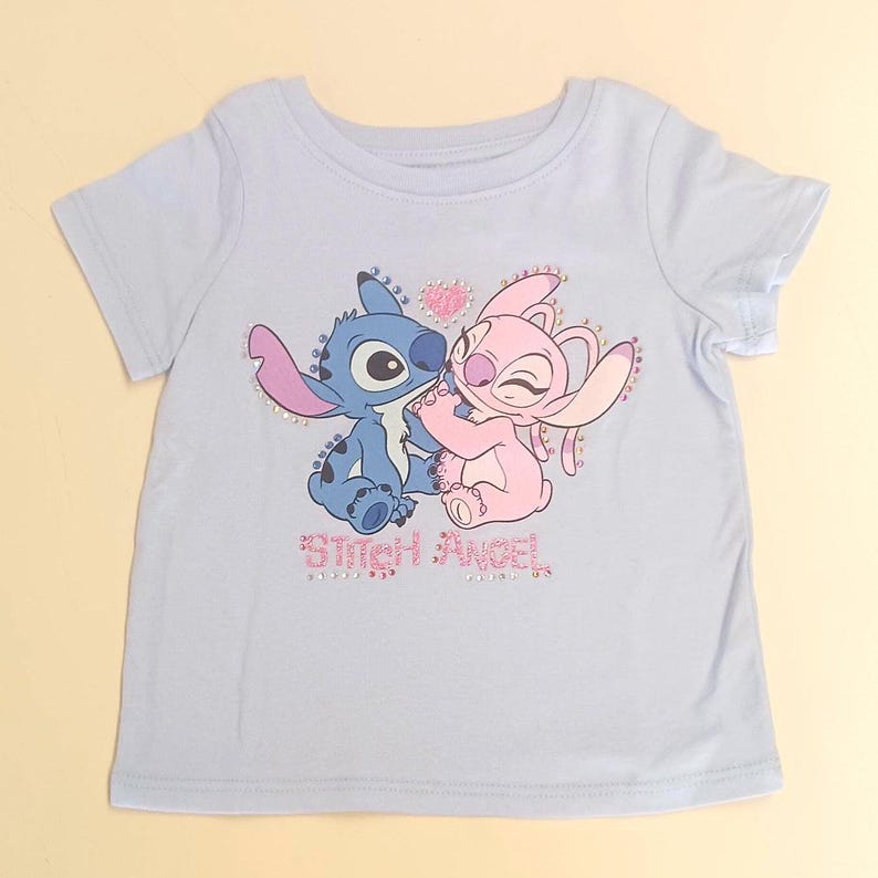 May include: A light blue t-shirt featuring a graphic of the characters Stitch and Angel embracing, with a pink heart above them. The text "Stitch Angel" is below the characters, with rhinestone accents.