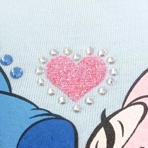 May include: Close-up of a light blue fabric with a pink glitter heart design. The heart is surrounded by clear rhinestones. Two blue rhinestones are visible on the left side of the image. Part of a cartoon character is visible.