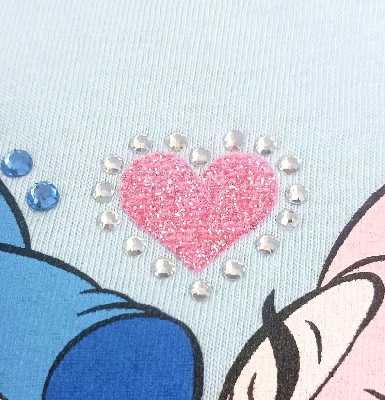 May include: Close-up of a light blue fabric with a pink glitter heart design. The heart is surrounded by clear rhinestones. Two blue rhinestones are visible on the left side of the image. Part of a cartoon character is visible.