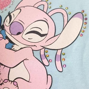 May include: Light blue fabric featuring a pink cartoon character with large ears and a purple nose. The character is decorated with colorful rhinestones and a pink glitter heart. The design is likely for children's apparel.