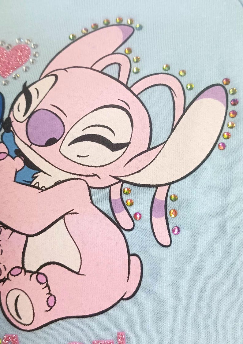 May include: Light blue fabric featuring a pink cartoon character with large ears and a purple nose. The character is decorated with colorful rhinestones and a pink glitter heart. The design is likely for children's apparel.