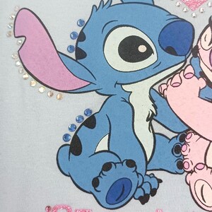 May include: Light blue graphic tee featuring the character Stitch from Lilo & Stitch. Stitch is blue with large ears and black spots. The text "Stitch & Angel" is in pink glitter, with rhinestones around the design.
