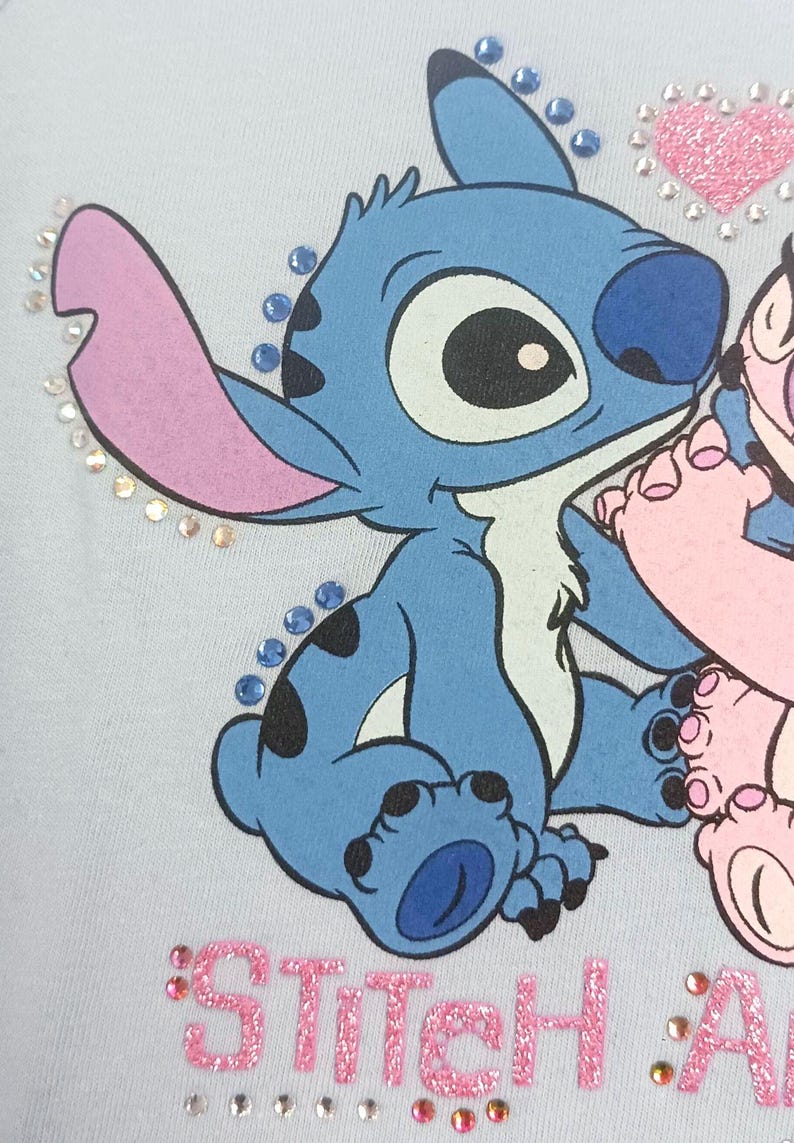 May include: Light blue graphic tee featuring the character Stitch from Lilo & Stitch. Stitch is blue with large ears and black spots. The text "Stitch & Angel" is in pink glitter, with rhinestones around the design.