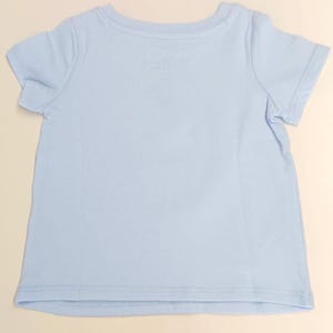 May include: A light blue short-sleeved t-shirt. The shirt has a crew neck and is made of a soft, lightweight fabric. The shirt is likely for a baby or toddler.