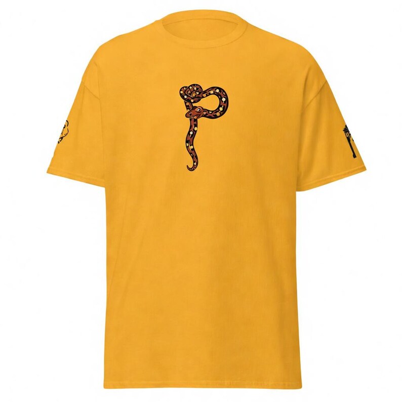 May include: Mustard yellow t-shirt with a detailed snake illustration in brown, red, and white. The snake is coiled in a stylized design. The shirt has short sleeves and a crew neck. The sleeves have a small graphic.