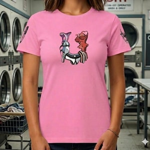 Op de afbeelding: Een roze t-shirt met een uniek grafisch ontwerp in de vorm van de letter "U". Het ontwerp bevat elementen van lingerie en abstracte vormen in grijstinten, bruin en wit. Het shirt heeft korte mouwen en een ronde hals.