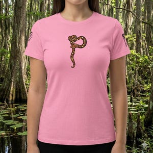 Könnte beinhalten: Ein pinkfarbenes T-Shirt mit einem braunen und gelben Schlangenmuster. Die Schlange ist oben aufgerollt und ihr Körper erstreckt sich nach unten. Das T-Shirt hat kurze Ärmel und einen Rundhalsausschnitt.