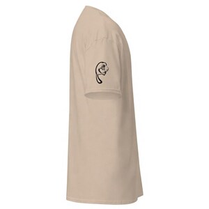 May include: A beige t-shirt with a black graphic on the left sleeve. The graphic is a stylized drawing of a face. The t-shirt is made of a soft material and has short sleeves.