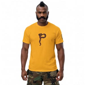 May include: Mustard yellow t-shirt with a brown and black snake design. The snake is coiled and has a long, curved tail. The shirt is short-sleeved and has a crew neck.