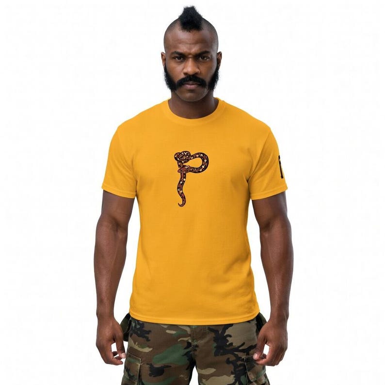 May include: Mustard yellow t-shirt with a brown and black snake design. The snake is coiled and has a long, curved tail. The shirt is short-sleeved and has a crew neck.