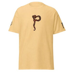 May include: Mustard-coloured t-shirt with a stylized snake design in brown, black, and red. The snake is in a curved, vertical position. The shirt has short sleeves and a crew neck.
