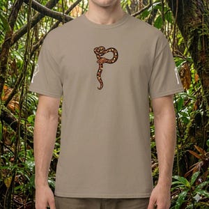 Könnte beinhalten: Ein beiges T-Shirt mit einer detaillierten Illustration einer braunen und beigefarbenen Schlange, die sich zu einer stilisierten Form windet. Das Schlangenmuster ist auf der Vorderseite des T-Shirts zentriert. Das T-Shirt hat kurze Ärmel und einen Rundhalsausschnitt.