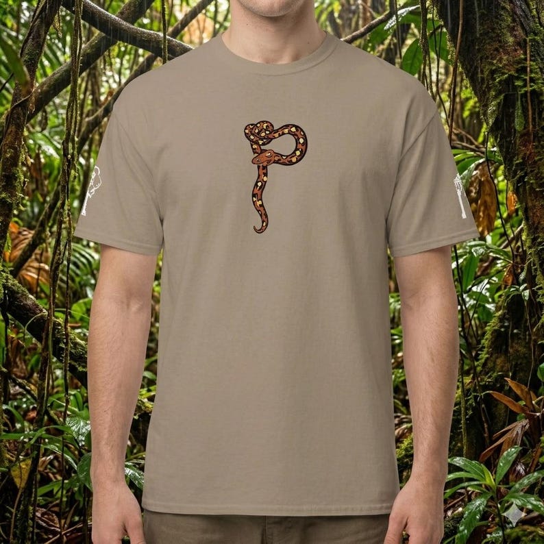 May include: A tan t-shirt with a detailed illustration of a brown and tan snake coiled into a stylized shape. The snake design is centered on the front of the shirt. The shirt has short sleeves and a crew neck.