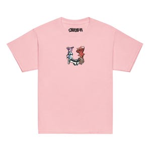 Op de afbeelding: Lichtroze t-shirt met een kleine afbeelding van twee cartoonfiguren en de letter "U" in het midden. Het woord "SIBNER" is in zwart boven de afbeelding gedrukt. Het shirt heeft korte mouwen en een ronde hals.