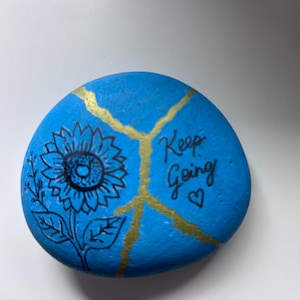May include: A smooth, round, blue painted rock featuring a black line drawing of a sunflower on the left side. A gold painted crack runs diagonally across the surface. The text "Keep Going" is written in black ink next to a small black heart.
