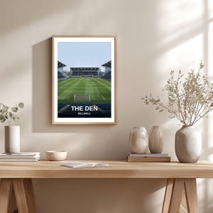 May include: Framed print of "The Den" stadium, featuring a green field, goal posts, and stadium seating under a blue sky. The print includes the text "THE DEN" and "MILLWALL" in white font. The artwork is displayed on a wooden table.