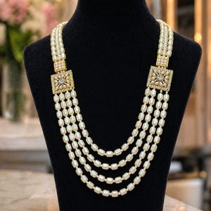 Peut inclure: Collier de perles à quatre rangs sur un mannequin noir. Le collier est composé de perles blanc cassé, reliées par des fermoirs carrés dorés avec des motifs détaillés. Les perles sont légèrement oblongues, pour un look classique.