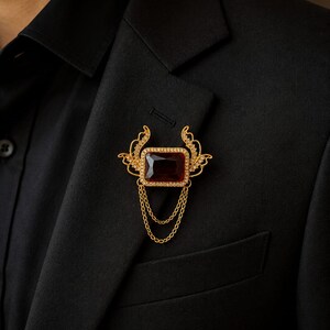 May include: A gold-toned brooch with a rectangular, dark red gemstone center, surrounded by smaller clear stones. The brooch has a decorative, wing-like design and two gold chains. It is pinned to a black blazer.