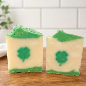 May include: Two rectangular bars of handmade soap featuring a cream-colored center and vibrant green layers on the top and bottom. Each bar displays a centered, green four-leaf clover design embedded within the cream-colored soap base.