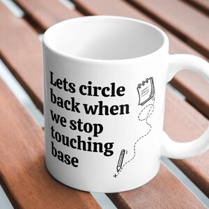 May include: White ceramic mug with black text that reads "Lets circle back when we stop touching base." A notepad and pencil graphic are also on the mug. The mug is on a wooden surface.