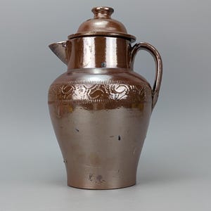May include: A brown glazed ceramic pitcher with a matching lid, a curved handle, and a pointed spout. The upper body features a decorative band with embossed swirling patterns and vertical indentations, finished with a glossy, reflective surface.