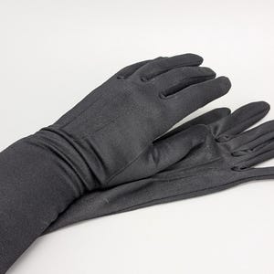 Peut inclure: Une paire de longs gants noirs unis fabriqués dans un tissu lisse et opaque. Les gants dépassent le poignet et présentent une coupe ajustée avec des coutures détaillées le long des doigts et du dos de la main, couvrant entièrement les mains.