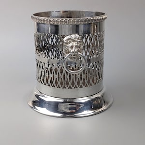 May include: A cylindrical silver-toned metal wine coaster featuring an intricate openwork lattice pattern, a decorative lion head emblem with a ring handle, and a smooth, flared base with a rope-textured rim along the top edge.