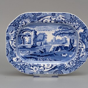 May include: An oval ceramic platter featuring a blue and white transferware pattern. The centre depicts a pastoral landscape with ruins, trees, and figures with livestock. The wide border is decorated with intricate floral and scroll motifs.