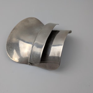 Può includere: Spilla astratta color argento con un design curvo e stratificato. La spilla presenta tre elementi distinti a forma di onda, creando un senso di profondità e movimento. Il metallo ha una finitura lucida, che riflette la luce e aggiunge alla sua estetica moderna.