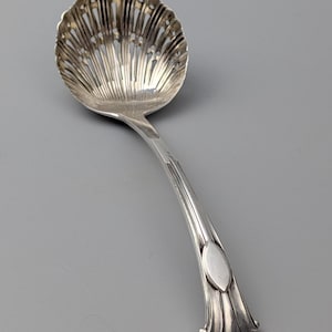 Op de afbeelding: Een zilverkleurige metalen serveerlepel met een kom in de vorm van een geschulpte schelp met decoratieve sleuven. Het handvat is gebogen en glad en eindigt in een uitlopende voet met een ovaal accent in het midden en verticale ribbels.