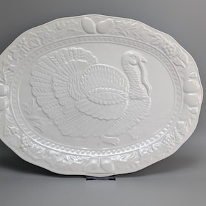 Puede incluir: Una bandeja de servicio ovalada de cerámica blanca con un diseño central de pavo en relieve. El borde está decorado con un patrón en relieve de varias frutas y hojas, creando un marco texturizado alrededor del motivo central del pavo.