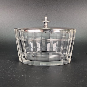 Könnte beinhalten: Eine klare Glasschale in konisch zulaufender zylindrischer Form mit einem gitterartigen Muster aus geätzten horizontalen und vertikalen Linien. Die Schale hat einen polierten silberfarbenen Metalldeckel mit einem kleinen, gestuften Knauf.