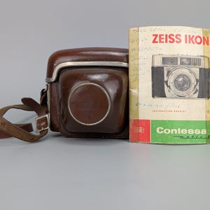 Op de afbeelding: Een bruine lederen cameratas met bijpassende schouderriem en een vintage Zeiss Ikon Contessa-matic E instructieboekje. Het boekje bevat een afbeelding van de camera, rode en groene accenten en handgeschreven notities op de omslag.
