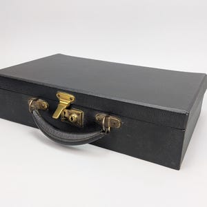 May include: A rectangular black carrying case with a textured finish, featuring a curved black handle and a central brass-colored metal latch mechanism with a keyhole. The case has a structured, boxy design with clean edges and a flat top surface.