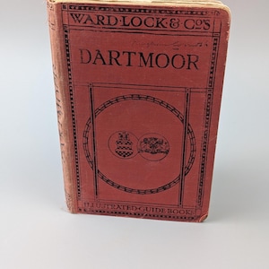 May include: A vintage red hardcover guidebook titled Ward Lock & Co's Dartmoor. The front cover features black text, decorative border lines, and two circular crest illustrations. The spine also displays the title Dartmoor in black lettering.