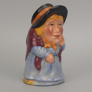 May include: A ceramic character jug shaped like a person wearing a black hat with an orange band, a maroon shawl, and a light blue dress. The figure has blonde hair, rosy cheeks, and a yellow neck scarf, depicted in a standing pose with one hand raised.
