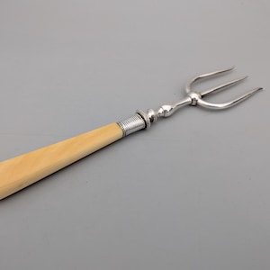 May include: A three-pronged silver fork featuring a smooth, cream-coloured handle. The handle connects to the metal tines via a decorative, ribbed silver ferrule. The fork has a polished metallic finish and a simple, elongated design.