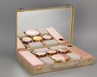 May include: A vintage vanity travel case featuring a hinged lid with an interior mirror. The case contains various rectangular and circular containers in shades of pale pink, white, and gold, organized within a structured, light-colored exterior frame.