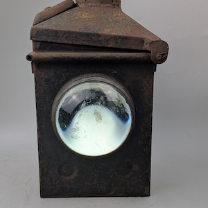 May include: A rectangular, dark brown, rusted metal lantern featuring a prominent circular glass lens on the front. The top section includes a small, domed chimney vent and a hinged lid. The overall surface shows significant oxidation and wear.