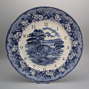 May include: A round ceramic wall clock featuring a blue and white transferware pattern. The center depicts a landscape with trees and a building, surrounded by black numerals from 1 to 12. The scalloped border displays floral motifs.