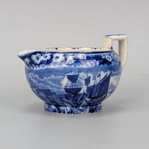 Puede incluir: Una jarrita de cerámica con un patrón de transferencia azul y blanco que representa barcos de vela en el agua. El borde está decorado con un diseño floral y la pieza incluye un pequeño pico vertedor y un asa cuadrada.