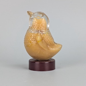 May include: A clear and amber-coloured glass bird figurine with a textured, pebbled surface on its body. The bird is perched atop a solid, dark brown cylindrical base. The glass features a smooth head and beak, contrasting with the textured chest.