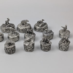 Könnte beinhalten: Eine Sammlung von zwölf kleinen, runden Schmuckdosen aus silberfarbenem Metall, jede mit einer einzigartigen, skulptierten Tier- oder Insektenfigur auf dem Deckel, darunter ein Eichhörnchen, ein Bär, ein Frosch, ein Schmetterling und Vögel.