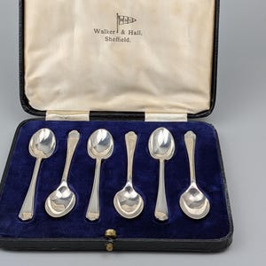 Può includere: Un set di sei cucchiai color argento disposti in una custodia nera con interno in velluto blu. L'interno del coperchio presenta la scritta Walker & Hall, Sheffield con un logo a bandiera. I cucchiai hanno manici lisci e lucidi.