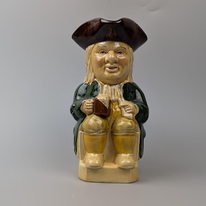 May include: A ceramic Toby jug figurine depicting a seated man in a tricorn hat and green coat. The jug has a brown hat, tan face, and yellow pants. The man is holding a brown object and a white scroll.