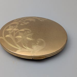 Op de afbeelding: Een goudkleurig, rond compactpoederdoosje met een geëtst bloemmotief op het deksel. Het compactpoederdoosje is gesloten en heeft een licht versleten uiterlijk, wat suggereert dat het vintage is. Het bloemmotief omvat gestileerde bloemen en bladeren.