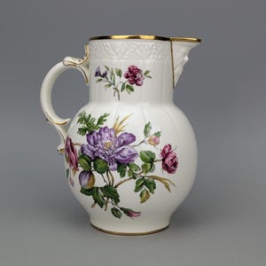 May include: A white porcelain pitcher featuring a hand-painted floral design with purple and pink flowers, green leaves, and gold trim along the rim and handle. The body has a textured upper section and a curved handle with gold accents.