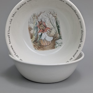 Op de afbeelding: Een witte keramische kom met een centrale illustratie van een konijn in een rode mantel, met een mand en een paraplu in een bosrijke omgeving. De binnenrand is voorzien van de tekst: Mrs. Rabbit took a basket and her umbrella and went through the wood to the bakers. Then old Mrs. Rabbit bought a loaf of brown bread.