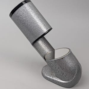 May include: A silver-toned metal desk lamp with a textured, hammered finish. The lamp features a cylindrical head with a black rim, connected by a slanted arm to a curved, oval-shaped base. The interior of the lamp head is white.