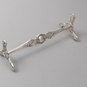 May include: A silver-colored metal knife rest featuring an elongated horizontal bar supported by two ornate, three-legged bases. The central section of the bar displays a circular medallion, while the entire structure is decorated with intricate scrollwork.